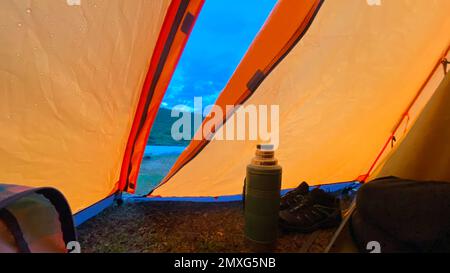 Thermos e scarpe da trekking si trovano all'uscita socchiusa della tenda in una serata piovosa sulle montagne Altai Foto Stock