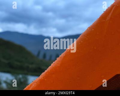 Le gocce di pioggia si trovano sulla tenda di una tenda turistica arancione sullo sfondo di una montagna e di un fiume. Foto Stock
