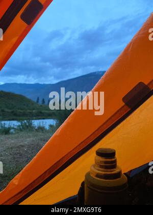 Le gocce di pioggia si trovano sulla tenda di una tenda turistica arancione con thermos sullo sfondo di una montagna e di un fiume. Telaio verticale. Foto Stock