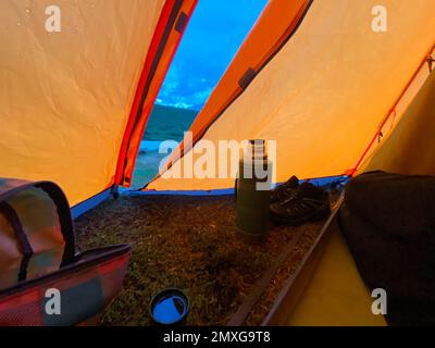 Le gocce di pioggia si trovano sulla tenda di una tenda turistica arancione con thermos di tè e scarpe sullo sfondo di una montagna e di un fiume in serata. Foto Stock