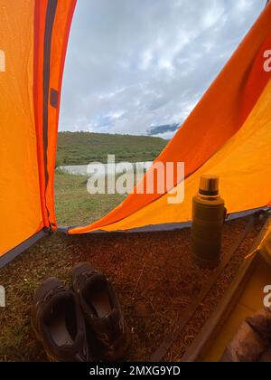 Una thermos e scarpe da trekking si trovano all'uscita semiaperta di una tenda in una serata piovosa sui monti Altai. telaio verticale Foto Stock