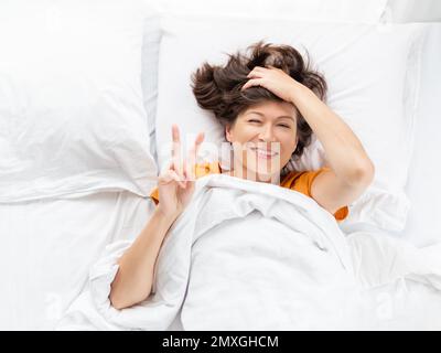 Vista dall'alto sulla donna addormentata che mostra il gesto della Vittoria a letto. La donna si allunga sdraiata sulla biancheria da letto bianca. Piacevole risveglio. Buon sonno. Foto Stock