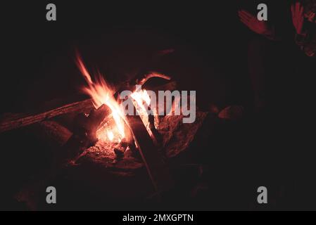 La ragazza riscalda le palme delle sue mani vicino alla fiamma calda di un fuoco con scintille e legna da ardere di notte. Foto Stock