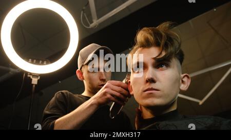 acconciatura e taglio dei capelli da uomo con regolacapelli in barbiere o parrucchiere. Servizio parrucchiere in un moderno barbiere in un fulmine chiave scuro Foto Stock