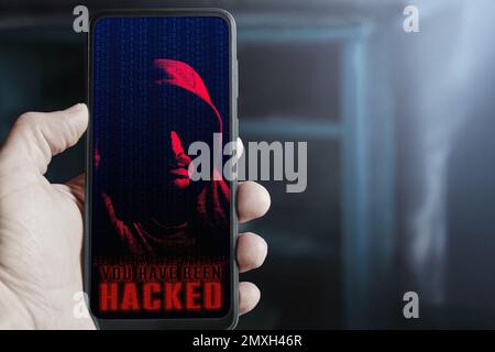 Mano maschile che tiene il telefono cellulare con l'avviso hacker e incappucciati cyber criminale sullo schermo. Concetto di sicurezza informatica, violazione dei dati dopo un attacco informatico. IO Foto Stock