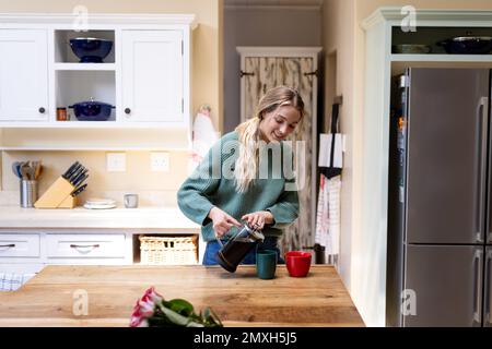 Sorridente biraciale bella giovane donna con capelli biondi versare caffè in tazze sul tavolo in cucina Foto Stock