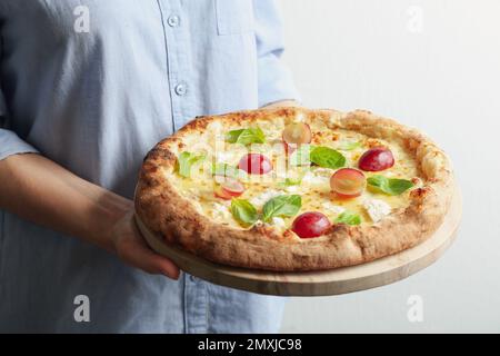 Donna che tiene deliziosa pizza al formaggio con uva e basilico su sfondo chiaro, primo piano Foto Stock