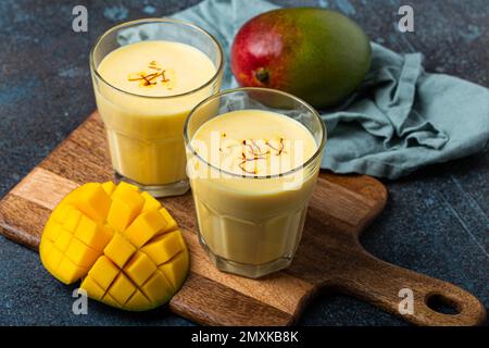 Sani Ayurveda indiano bere mango lassi in due bicchieri su tavolo rustico in cemento con mango fresco tagliato maturo, bevanda mista gialla fatta di mango fru Foto Stock