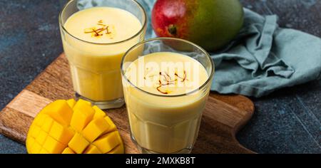 Sani Ayurveda indiano bere mango lassi in due bicchieri su tavolo rustico in cemento con mango fresco tagliato maturo, bevanda mista gialla fatta di mango fru Foto Stock