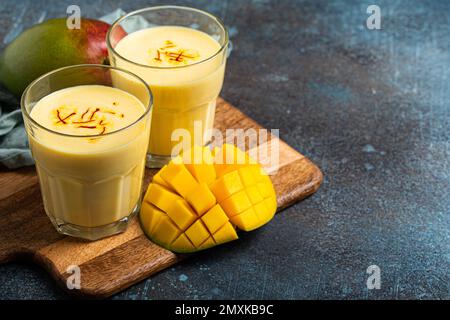 Sani Ayurveda indiano bere mango lassi in due bicchieri su tavolo rustico in cemento con mango fresco tagliato maturo, bevanda mista gialla fatta di mango fru Foto Stock