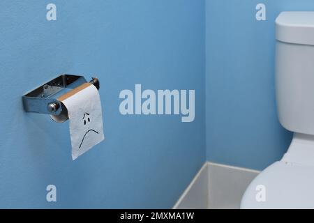 Un'ampia scena in una toilette di una faccia triste sull'ultimo pezzo di carta igienica/tessuto su un rotolo di carta igienica di cartone marrone, su uno sfondo di parete blu chiaro Foto Stock