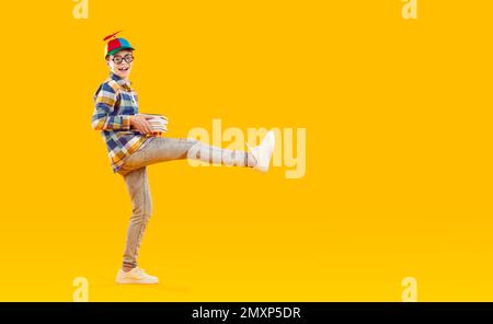 Ragazzo di scuola in cappellino divertente e bicchieri che tengono libri e in piedi su sfondo giallo copia spazio Foto Stock