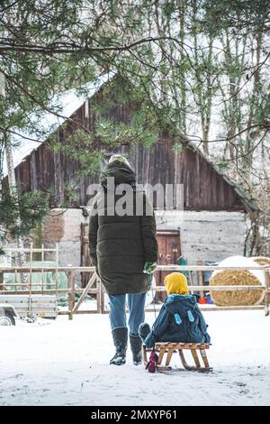 Madre o ragazza o donna che camminano in campagna in inverno con bambino nella slitta e bianco paesaggio invernale, casa di legno, fattoria, balla di fieno Foto Stock