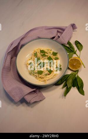 Pasta cremosa con condimento al basilico. Su fondo in marmo bianco con tovaglia lilla, limone e basilico fresco. Preso dall'alto. Foto Stock