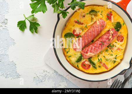 Omelette o frittata con due salsicce, formaggio fuso, pomodori ciliegini, broccoli e prezzemolo su padella rossa o teglia di ferro su fondo bianco di cemento vecchio Foto Stock