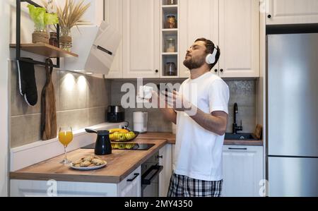 Uomo in cuffia in piedi in cucina domestica bere caffè e ascoltare musica godendo del tempo di mattina. Foto Stock