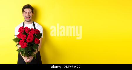 Venditore in negozio di fiori che indossa grembiule nero, dando bouquet di rose e sorridente, in piedi su sfondo giallo Foto Stock