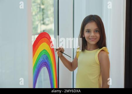 Piccola ragazza disegno arcobaleno sulla finestra all'interno. Concetto di soggiorno a casa Foto Stock