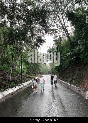 madre latina che cammina con i suoi figli su una strada solitaria in una giornata di pioggia, circondata dalla natura. Foto Stock