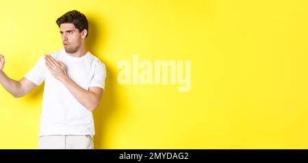 Uomo che mostra abilità kung-fu, arti marziali movimento ninja, in piedi in bianco t-shirt pronto a combattere, in piedi su sfondo giallo Foto Stock