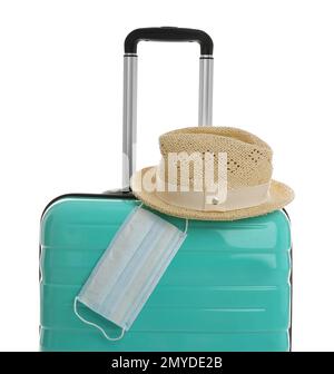 Elegante valigia turchese, cappello e maschera protettiva su sfondo bianco. Viaggiare durante la pandemia di coronavirus Foto Stock