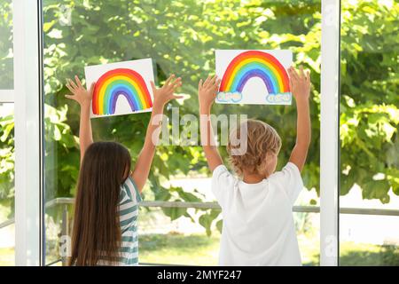 Bambini piccoli che tengono dipinti arcobaleno vicino alla finestra al coperto, vista sul retro. Concetto di soggiorno a casa Foto Stock