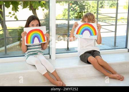 Bambini piccoli che tengono dipinti arcobaleno vicino alla finestra al coperto. Concetto di soggiorno a casa Foto Stock