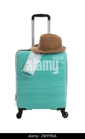 Elegante valigia turchese, cappello e maschera protettiva su sfondo bianco. Viaggiare durante la pandemia di coronavirus Foto Stock