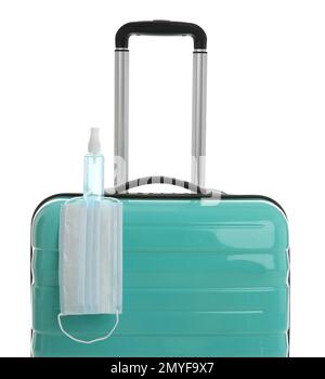 Elegante valigia turchese, spray antisettico e maschera protettiva su sfondo bianco. Viaggiare durante la pandemia di coronavirus Foto Stock