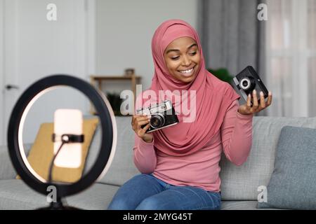 Entusiasta signora musulmana nero riprese video per blog, facendo recensione su fotocamere retro, seduto a casa Foto Stock