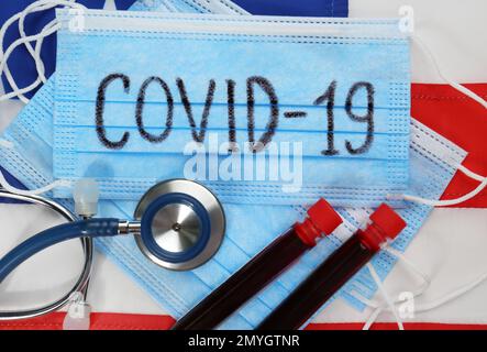 COVID-19 scritto su maschera di protezione, provette con sangue, stetoscopio e bandiera americana, piatto. Pandemia di coronavirus negli Stati Uniti Foto Stock