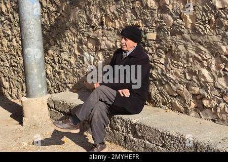Un uomo anziano guardò le attività della sua città dell'Iraq settentrionale dal suo persico su una panca di cemento appoggiata contro un muro di pietra. Foto Stock