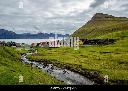 Villaggio di Gjogv visto da attraverso un ruscello che guarda fuori al fiordo con campi erbosi e montagna su Eysturoy, Isole Faroe Foto Stock