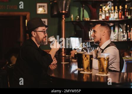 l'uomo si diverte a chiacchierare con un barista al bancone del bar mentre beve birra da un bicchiere in un pub Foto Stock