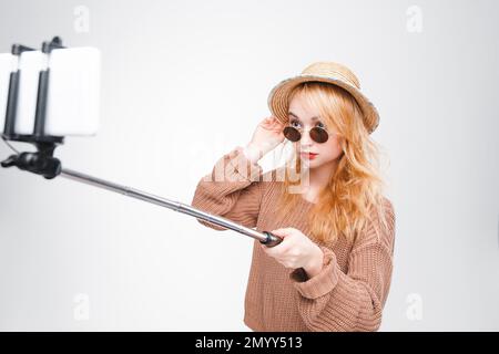 Ragazza giovane in occhiali da sole e cappello di paglia scatta foto con bastone selfie. Ragazza Hippster alla moda che si diverte. Blogger di moda. Sfondo grigio con copia Foto Stock