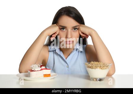 Donna dubbia scelta tra yogurt con muesli e torta a tavola su sfondo bianco Foto Stock