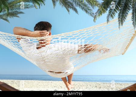 Uomo che si rilassa in amaca sotto le foglie di palma verde sulla spiaggia illuminata dal sole Foto Stock