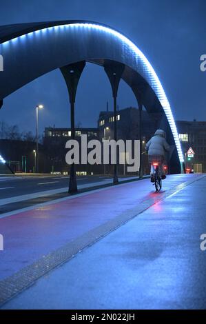 Donna con giacca grigia in bicicletta sotto la pioggia sul ponte autostradale illuminato di Heerdt a Duesseldorf, Germania Foto Stock