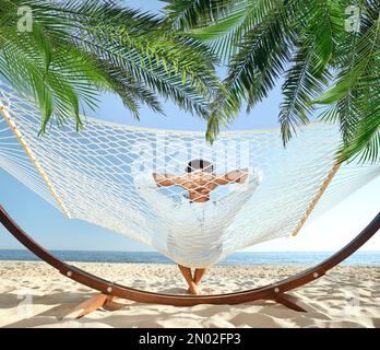 Uomo che si rilassa in amaca sotto le foglie di palma verde sulla spiaggia illuminata dal sole Foto Stock