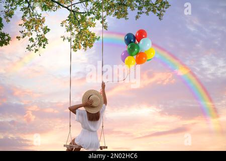 Il mondo dei sogni. Giovane donna con palloncini luminosi che oscillano, arcobaleno nel cielo del tramonto sullo sfondo Foto Stock