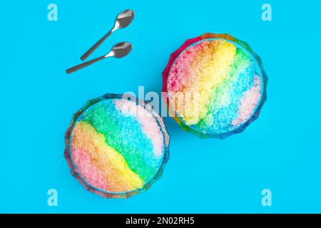 Rasatore di ghiaccio arcobaleno in ciotole di vetro da dessert e cucchiai su sfondo azzurro, piatto Foto Stock