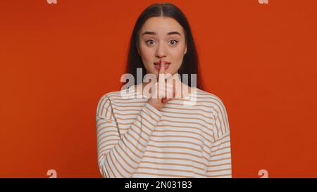 Shh Be Silenzio per favore. Ritratto di donna millenaria di 20 anni preme indice dito a labbra rende silenzio gesto segno non dice segreto. Giovane bella ragazza che posa su sfondo rosso studio Foto Stock