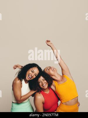 Tre giovani donne che ridono felicemente in uno studio che indossa abiti sportivi. Gruppo di amici femminili che celebrano la loro forma, sano Foto Stock