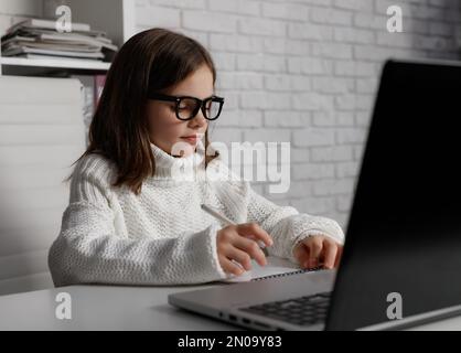 Bambina in occhiali che usa il notebook a casa. Capretto che fa i compiti. Brunetta caucasica al tavolo bianco. Concetto di formazione online Foto Stock