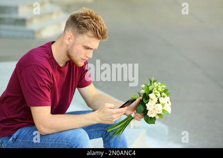 Giovane uomo con bouquet di fiori e telefono in attesa della sua ragazza all'aperto Foto Stock