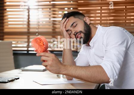 Uomo con ventola portatile che soffre di calore sul luogo di lavoro. Stagione estiva Foto Stock