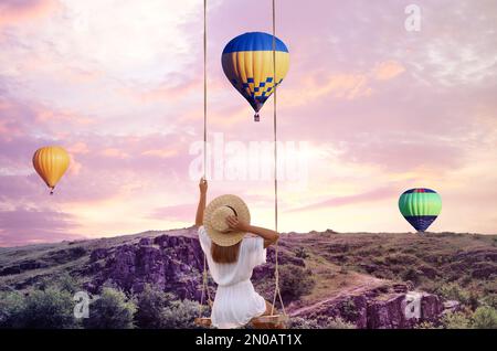 Il mondo dei sogni. Giovane donna che oscilla sulle montagne, mongolfiere nel cielo del tramonto sullo sfondo Foto Stock