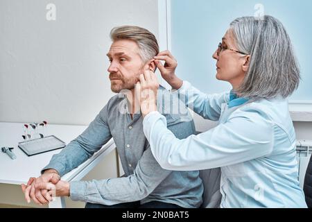 Medico di sesso femminile esperto che inserisce l'apparecchio acustico intraauricolare nell'orecchio del paziente durante la consultazione in clinica audiologica. Apparecchi acustici ITE e ITC Foto Stock