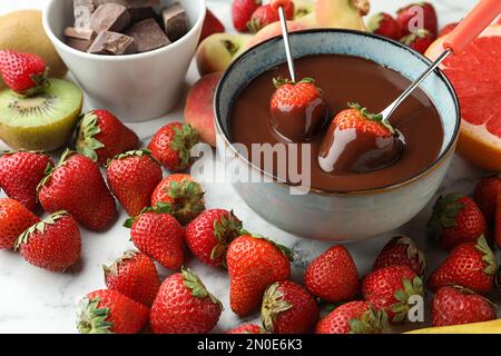 Forchette di fonduta con fragole in una ciotola di cioccolato fuso circondata da altri frutti sul tavolo Foto Stock