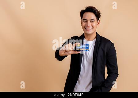 Felice uomo d'affari asiatico sorridente che tiene una carta di credito durante la presentazione. Uomo gioioso alla moda in camicia bianca e vestito su sfondo di colore beige - con spazio copia Foto Stock